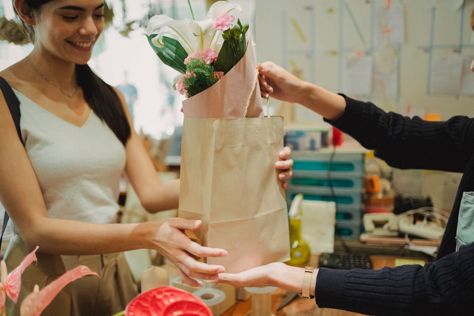 A woman with long dark hair, dressed in a white sleeveless top, is smiling as she receives a bouquet of fresh flowers from another person in a floral shop environment. The bouquet features a variety o