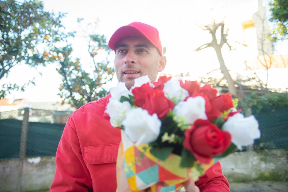 A delivery person wearing a red cap and red jacket holds a vibrant floral bouquet outdoors, with red and white roses and greenery arranged in a colorful wrapping paper. The background features a garde