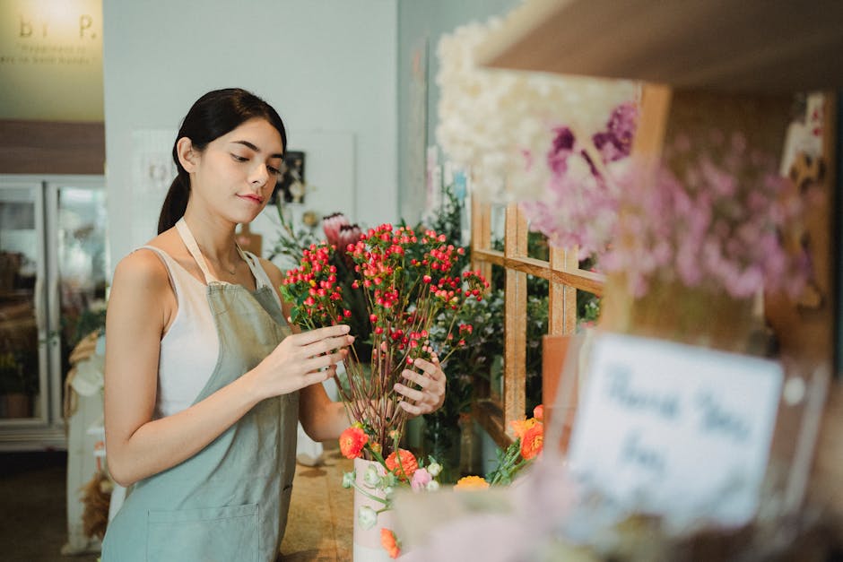 A young female florist with dark hair tied back, wearing a white sleeveless top and a beige apron, arranging a bouquet of vibrant pink and red hypericum berries with green stems in a flower shop. The 