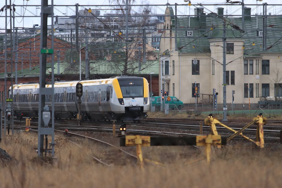 A modern passenger train with a sleek silver and yellow exterior traveling along railway tracks near West Dulwich station SE21. The train is set against a background of residential buildings with gree