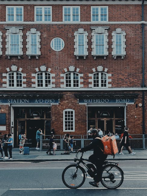 A person riding a bicycle on the street in front of a brick building with large arched windows and a sign that reads 'Station A.D. 1910 Waterloo Road,' while other pedestrians walk along the sidewalk.