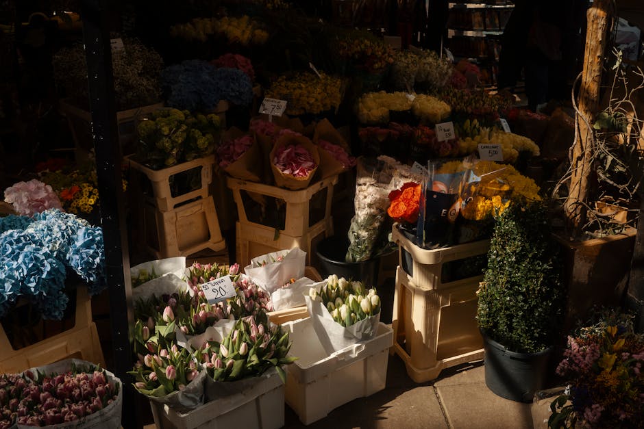 A variety of fresh flowers arranged in bouquets and baskets at a flower market, including pink tulips in a white paper wrap, blue hydrangeas, yellow chrysanthemums, and orange roses. These flowers are